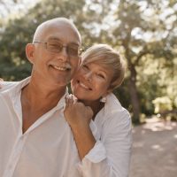 Smiling old man with grey hair and mustache in glasses and light shirt hugging with blonde woman in white clothes in park..